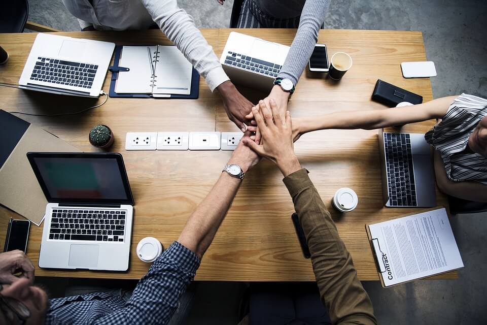 An image of the stacked hands of five people from diverse backgrounds with computers and other work items, representing the benefits of a diverse workforce and how avoiding WV employment discrimination can be achieved.