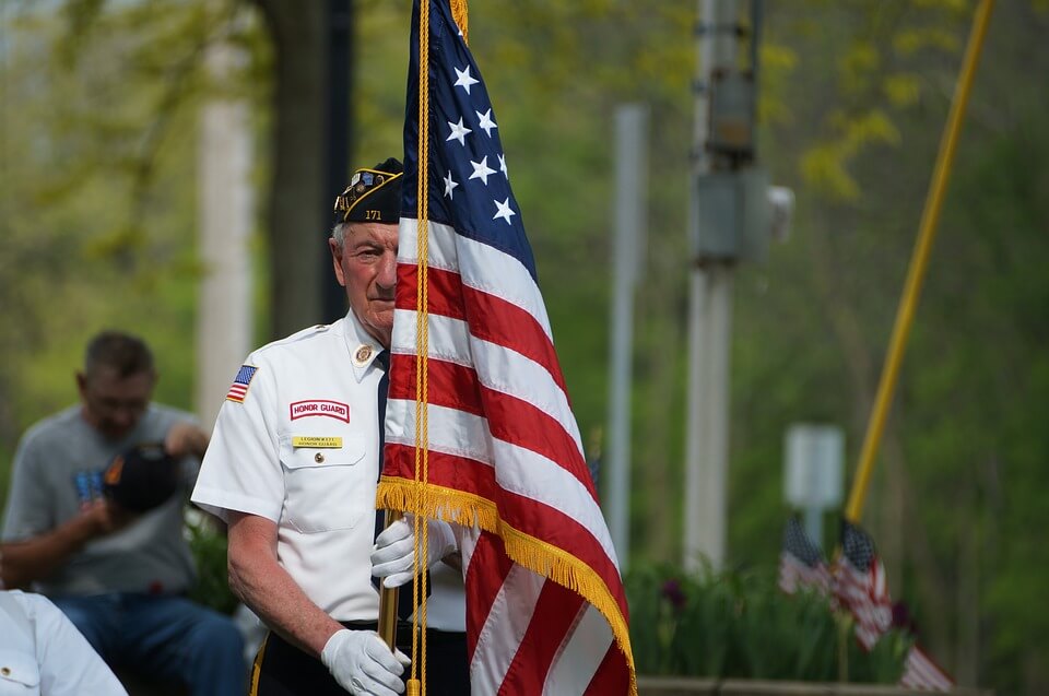 An image of an elderly man in a military uniform holding an American flag, representing those who have served in the US military and the veterans’ aid and attendance benefit that might be available to them with the help of an experienced estate planning attorney.